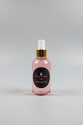 A bottle of perfume body mist with a gold spray nozzle and a black circular label reading 'Perfume Body Mist SHOPFUELTRAVEL.COM', placed on a light wooden table in a kitchen or storage room setting.
